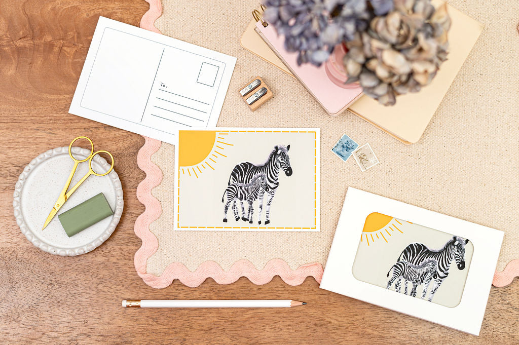 A6 Postcard with a Zebra and it's Baby, with a yellow sun in the background. The Card is on a desk, surrounded by Stationery.