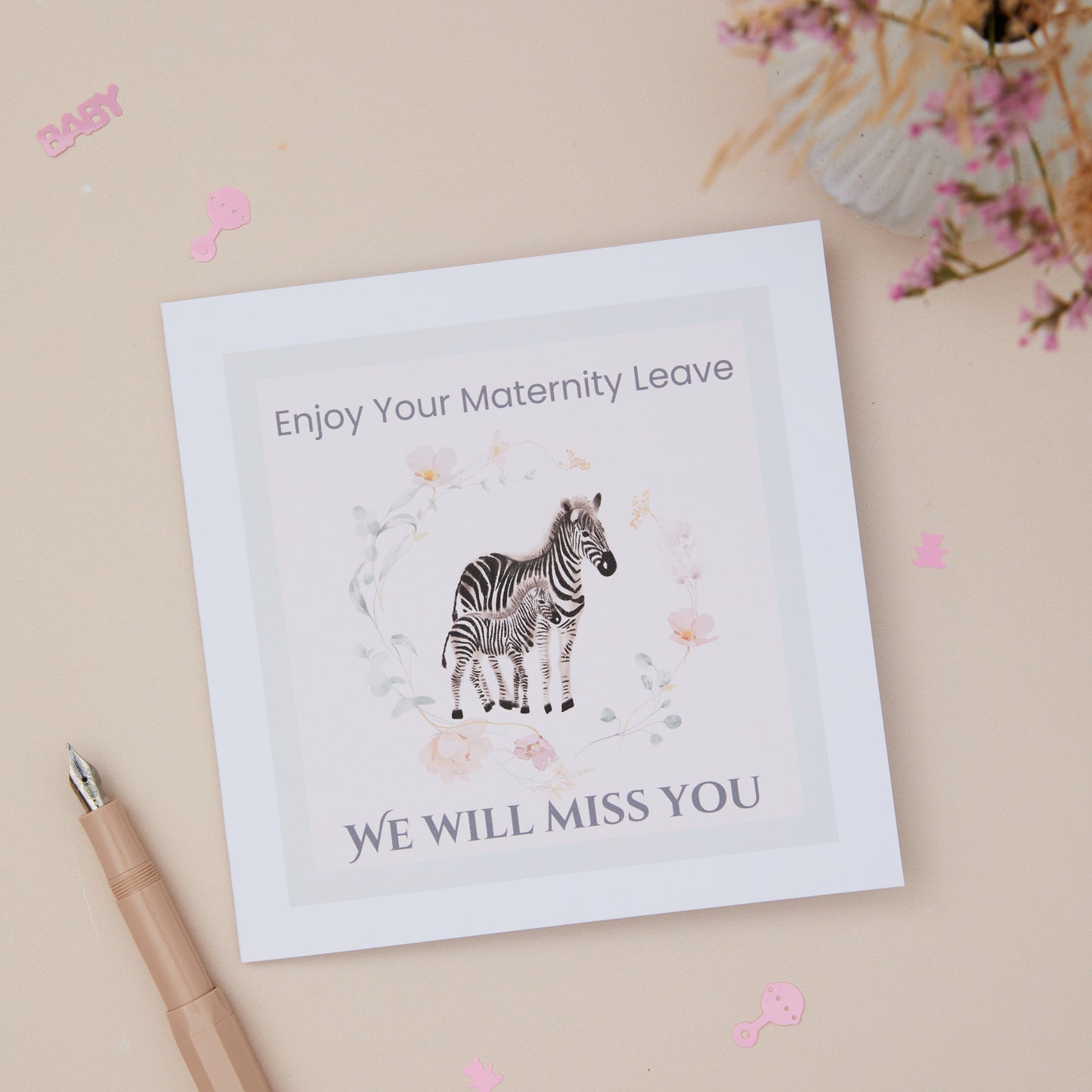 A greeting card with a zebra and its young, surrounded by floral elements, with text that reads 'Enjoy Your Maternity Leave - WE WILL MISS YOU'. There's a pen and pink decorative items at the bottom.