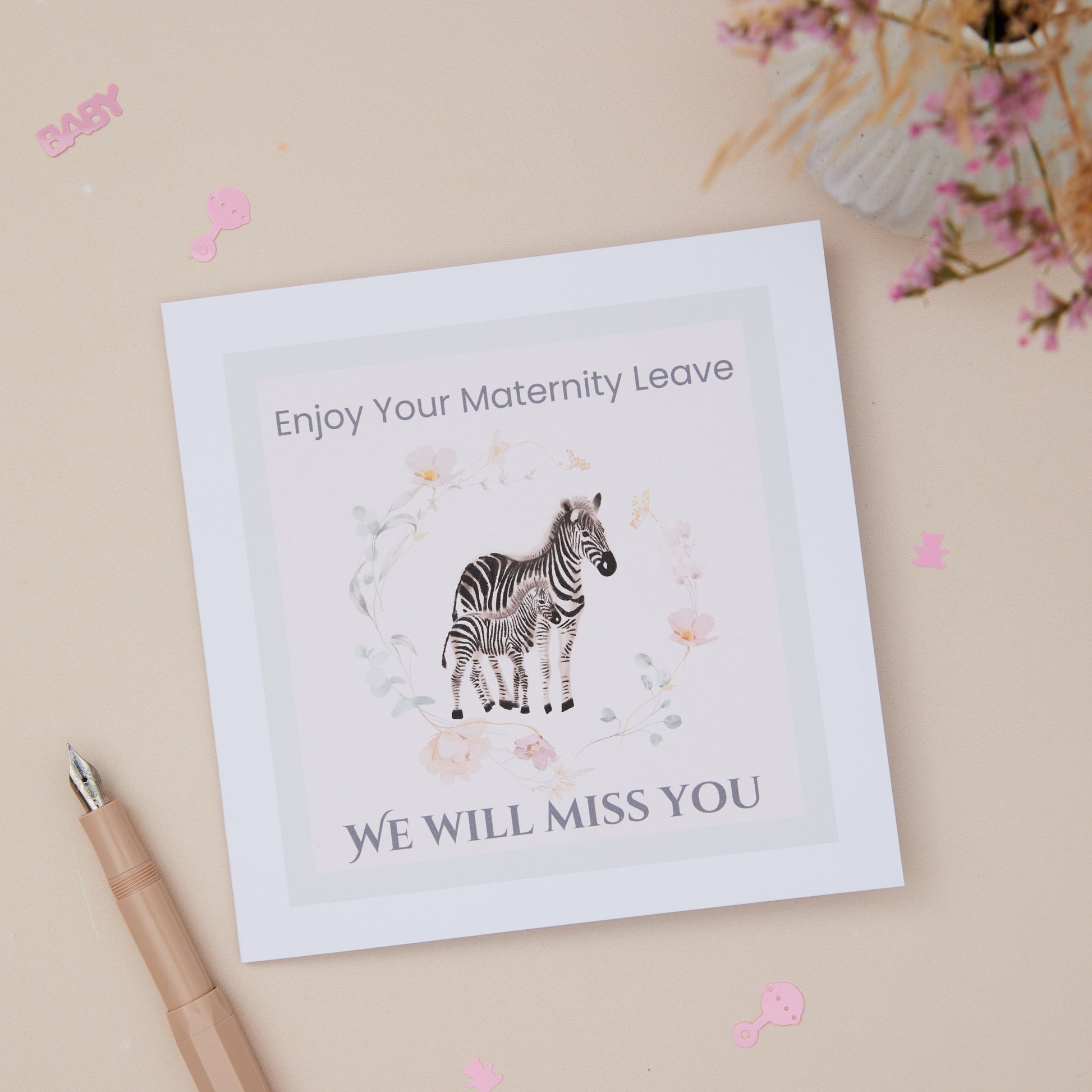 A greeting card with a zebra and its young, surrounded by floral elements, with text that reads 'Enjoy Your Maternity Leave - WE WILL MISS YOU'. There's a pen and pink decorative items at the bottom.